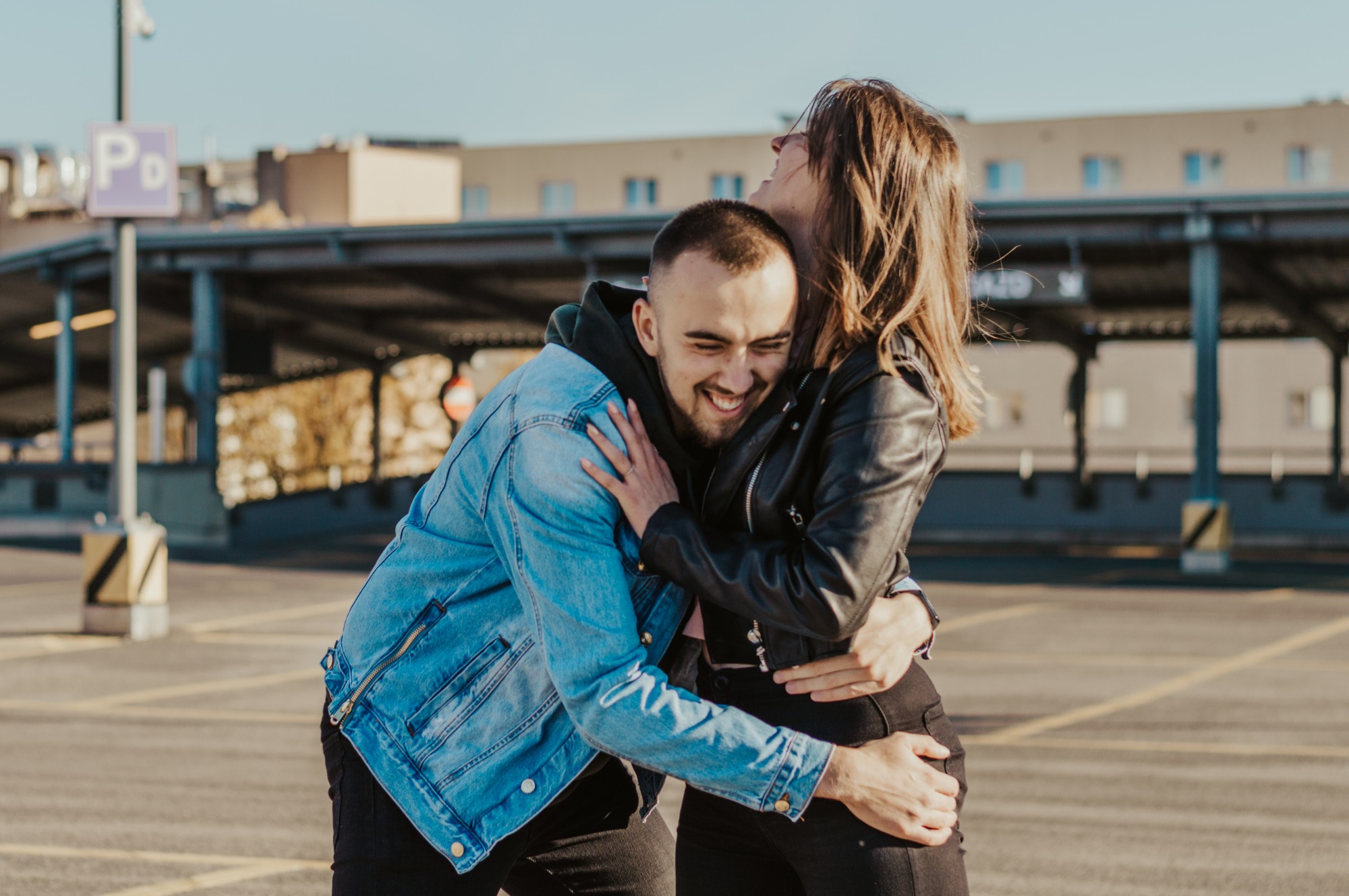 Uśmiechnięta para, mężczyzna w jeansowej kurtce i kobieta w czarnej skórzanej kurtce, obejmują się na parkingu wielopoziomowym w słoneczny dzień.