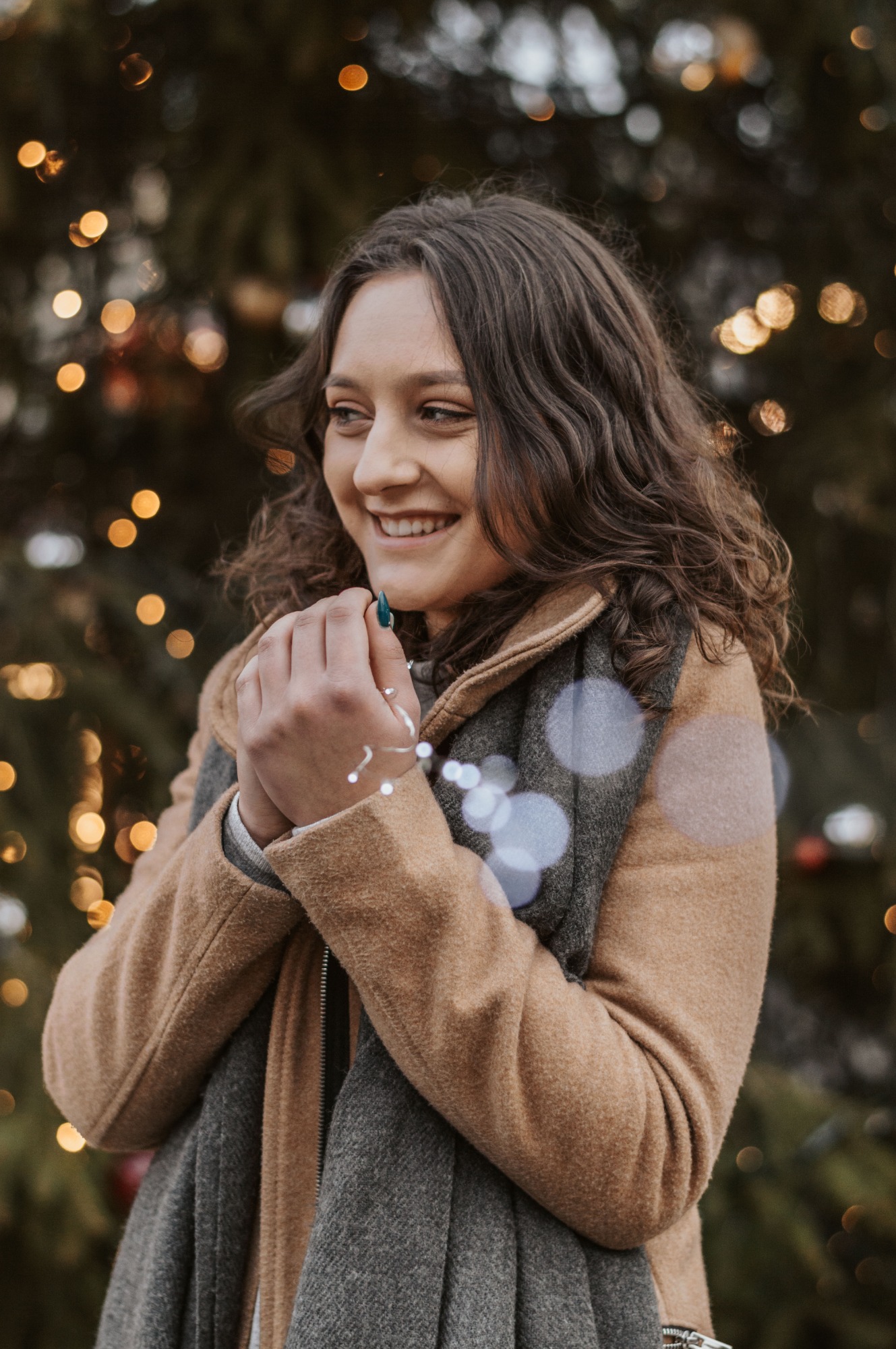 Uśmiechnięta kobieta w płaszczu, trzymająca w dłoniach światełka na tle choinki z lampkami.