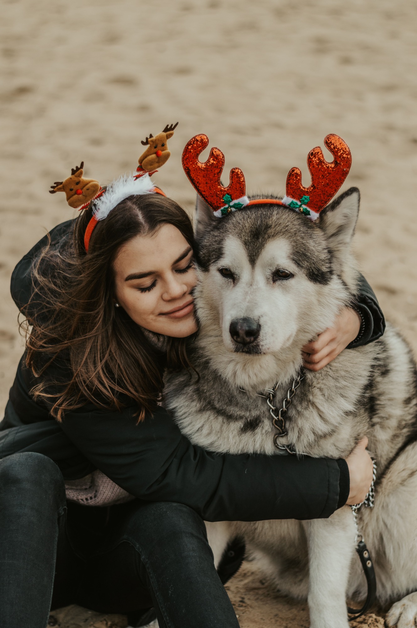 Młoda kobieta w czarnej kurtce, z zamkniętymi oczami, przytula dużego psa rasy Alaskan Malamute w świątecznych opaskach renifera na piaszczystym tle.