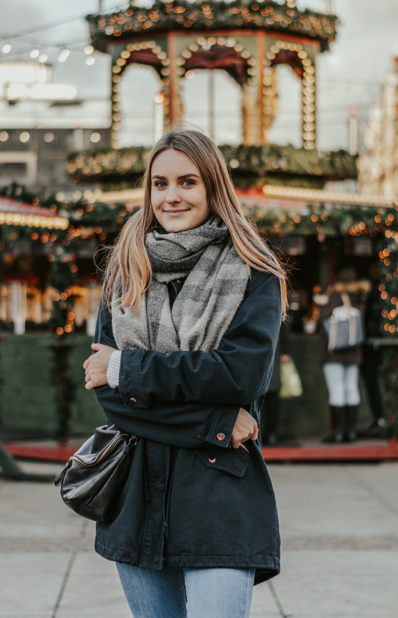 Uśmiechnięta blondynka w szarym szaliku i granatowej kurtce, pozuje na tle jarmarku bożonarodzeniowego z rozświetlonymi straganami i dekoracjami.