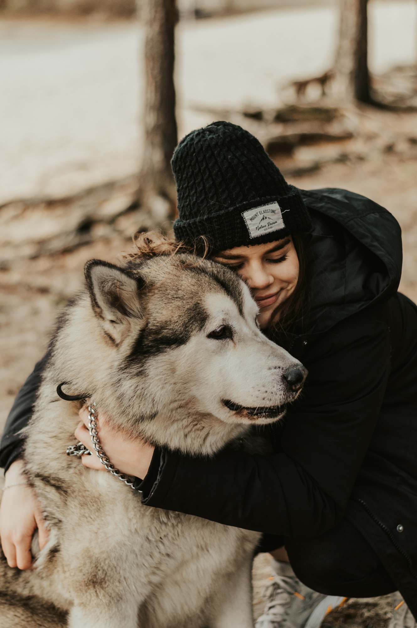 Portret młodej kobiety w czapce, czule obejmującej dużego psa rasy Alaskan Malamute na tle drzew.
