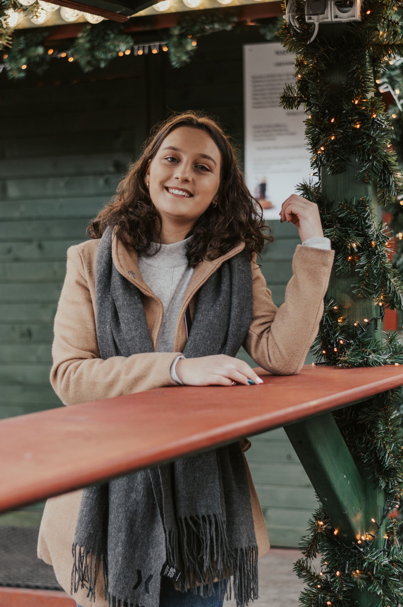 Uśmiechnięta kobieta w płaszczu i szaliku, opierająca się o czerwony blat przyozdobiony świątecznymi girlandami z lampkami, na tle zielonej drewnianej ściany.