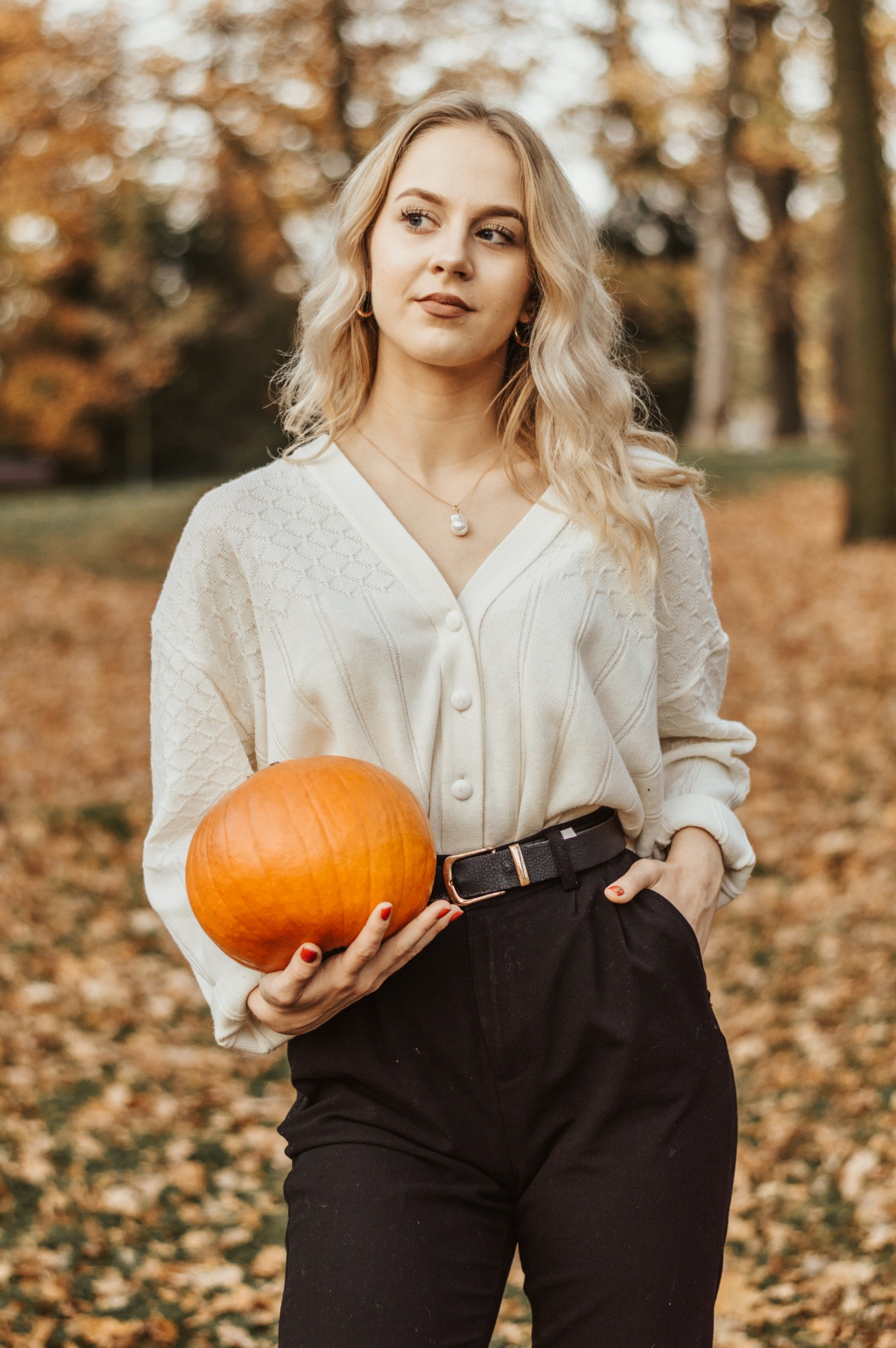 Portret młodej kobiety z blond włosami, trzymającej dynię na tle jesiennego parku, ubrana w kremowy kardigan i czarne spodnie.