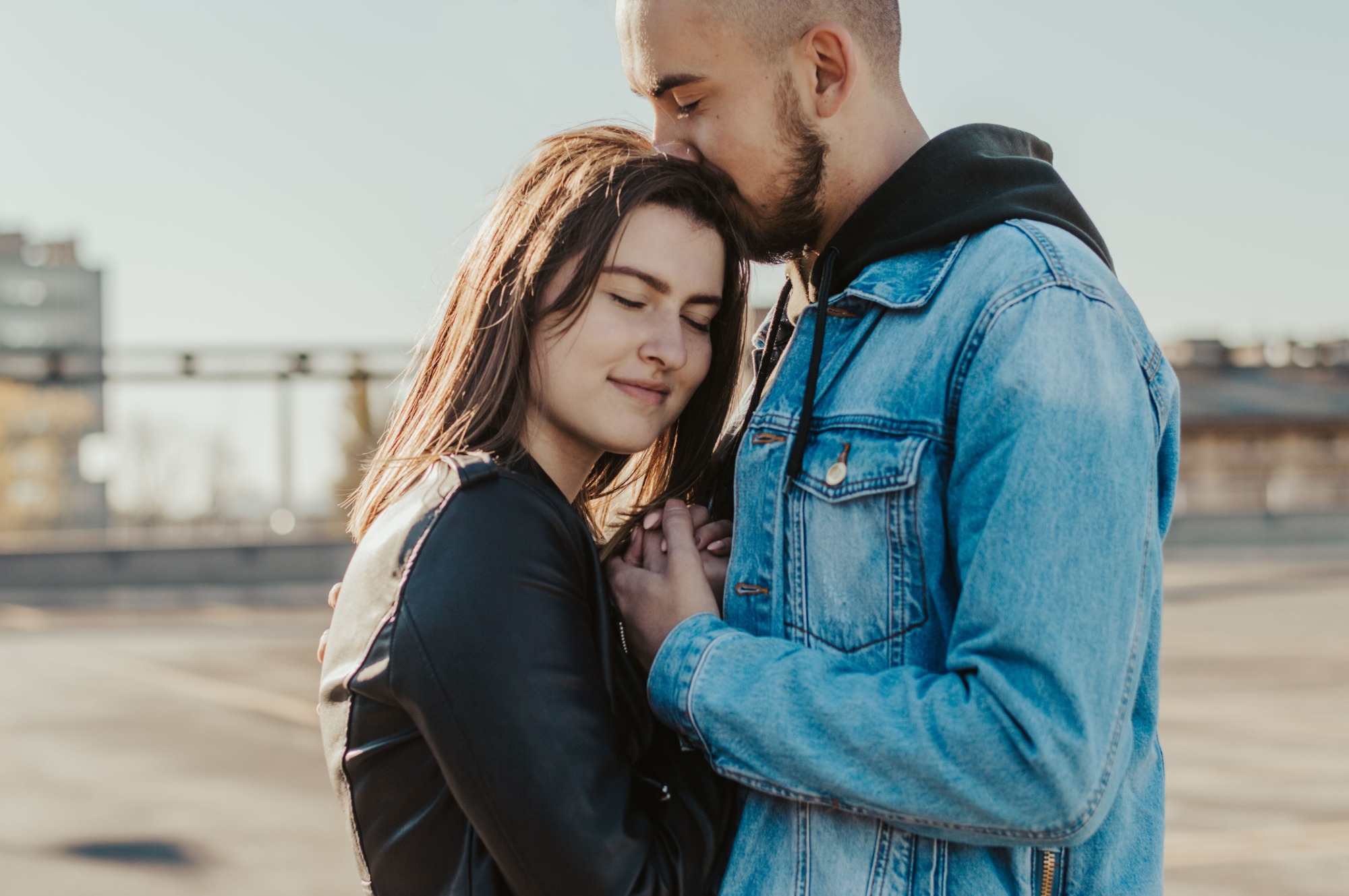 Młoda para: mężczyzna całuje kobietę w czoło, trzymając się za ręce na tle miejskiego krajobrazu, ona ubrana w skórzaną kurtkę, on w jeansową, oboje z zamkniętymi oczami.