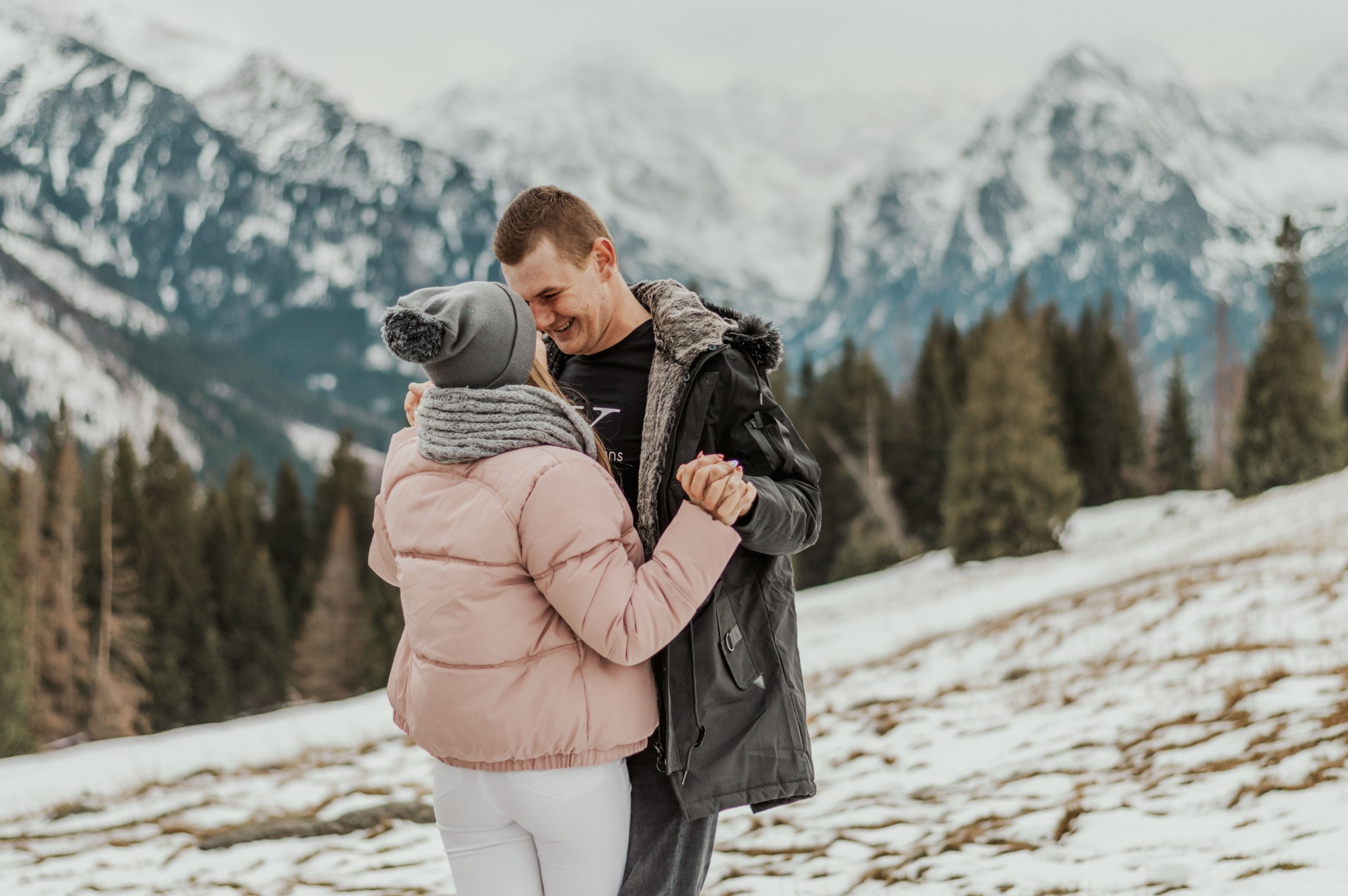 Para trzymająca się za ręce na tle ośnieżonych gór, kobieta w różowej kurtce i szarym szaliku, mężczyzna w czarnej kurtce z futrzanym kołnierzem, oboje uśmiechnięci.