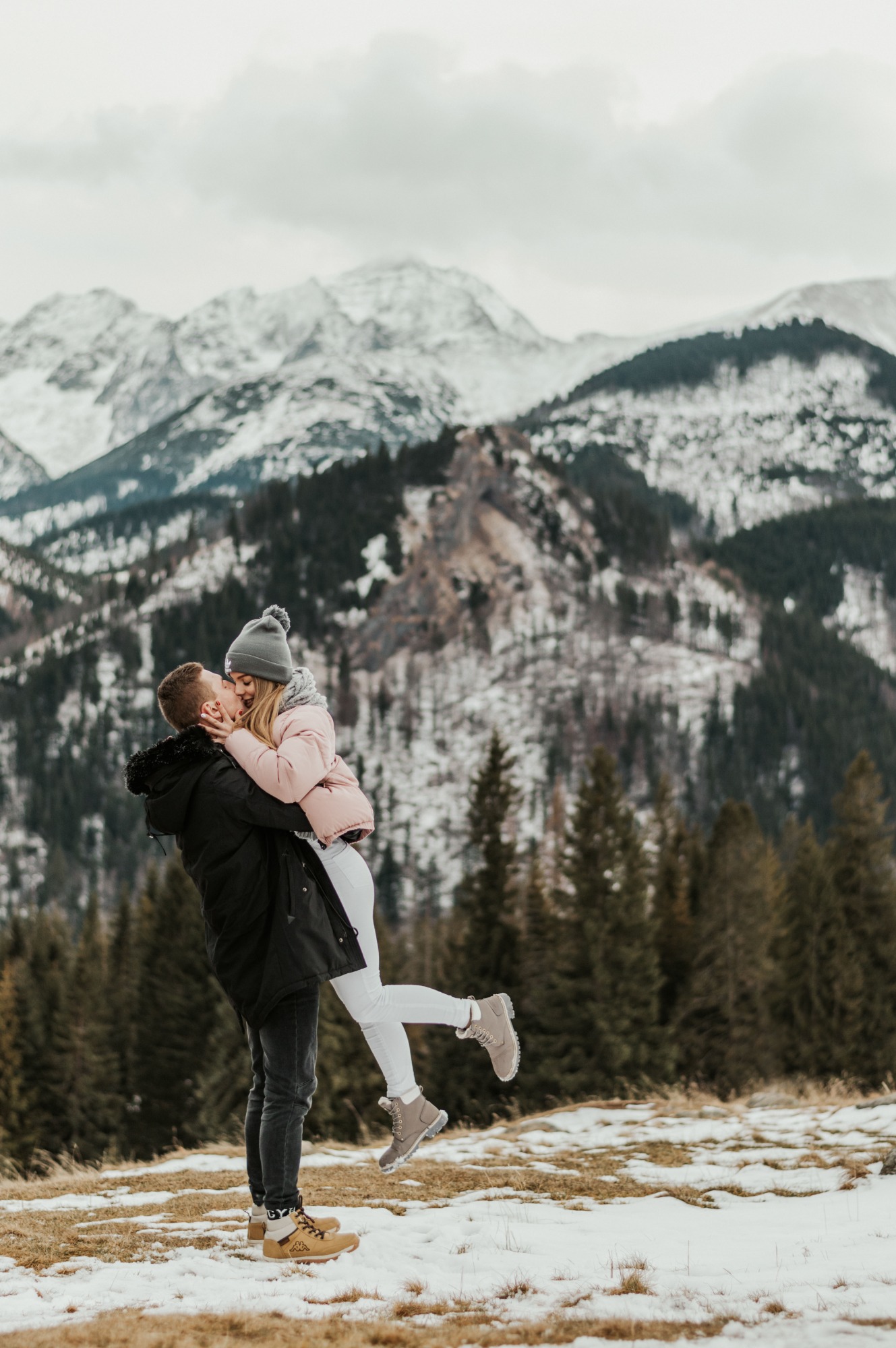 Mężczyzna trzyma na rękach kobietę, całując ją na tle gór i lasu w zimowej scenerii.