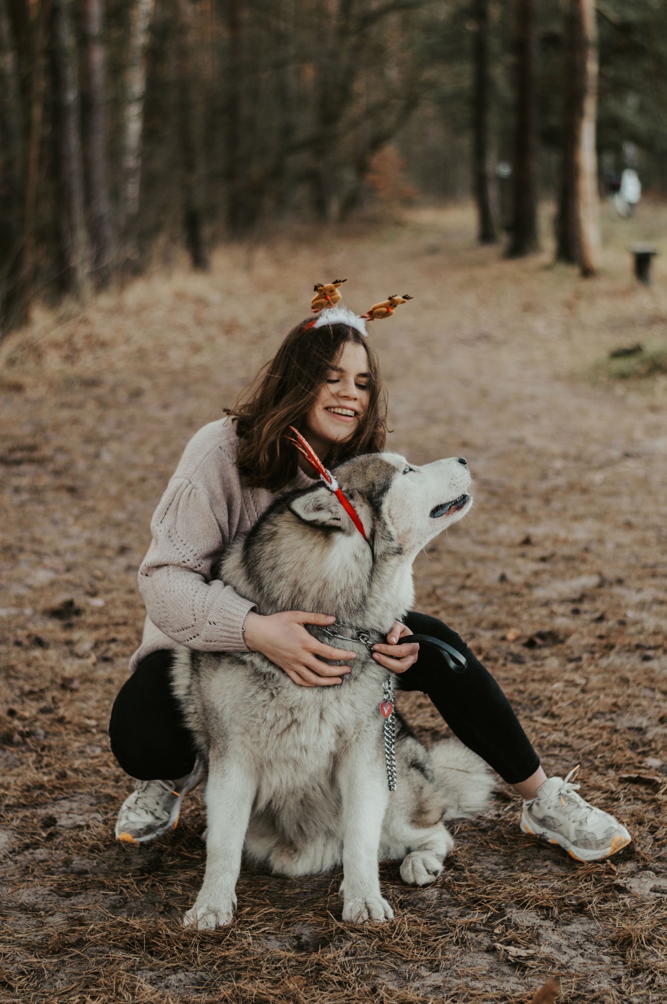 Uśmiechnięta brunetka w opasce z reniferami obejmuje dużego psa rasy Alaskan Malamute z czerwoną ozdobą na obroży w leśnej scenerii.