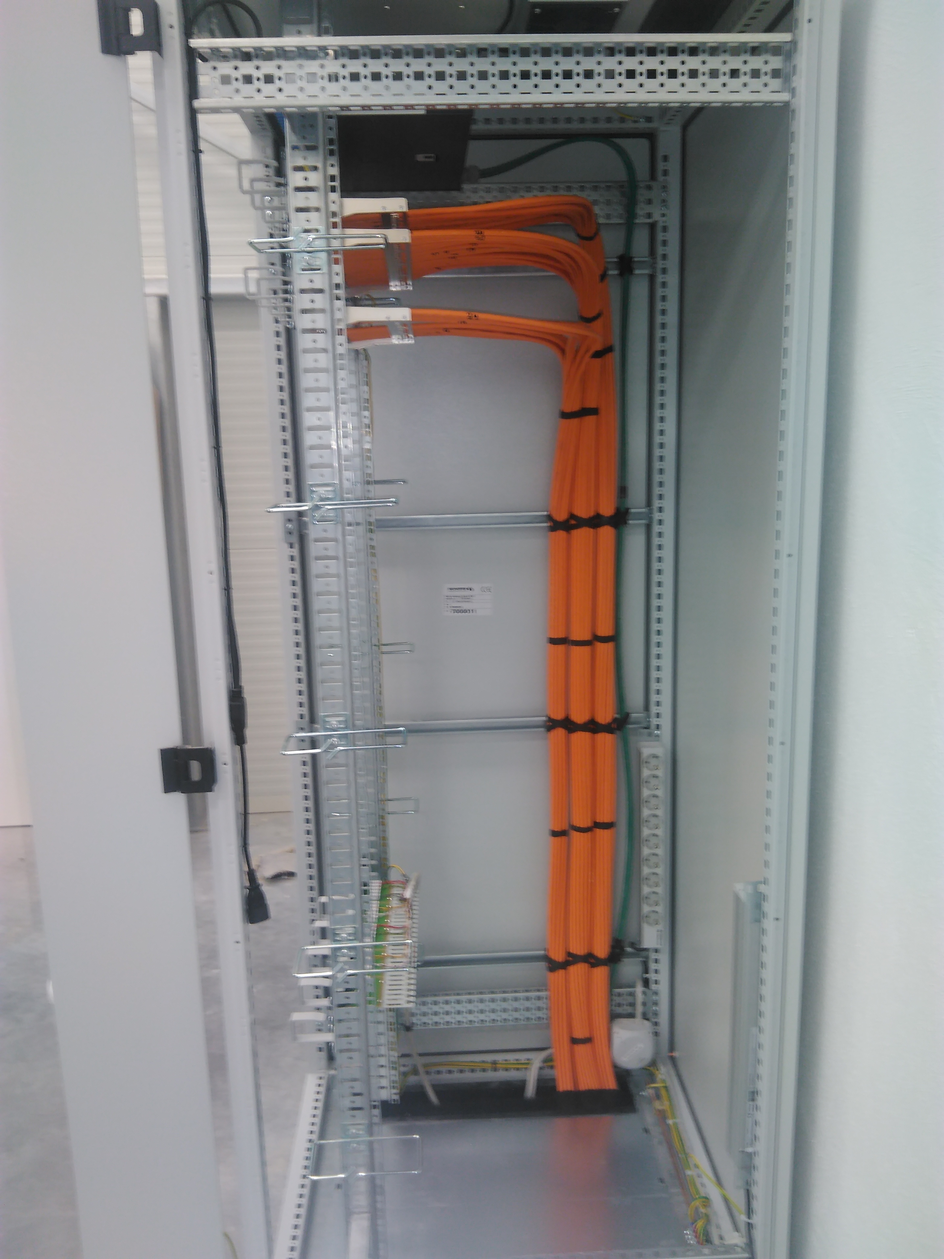 Wnętrze szafy rack z uporządkowanymi pomarańczowymi kablami, zabezpieczonymi czarnymi opaskami, widoczne szyny montażowe i listwa z gniazdkami.