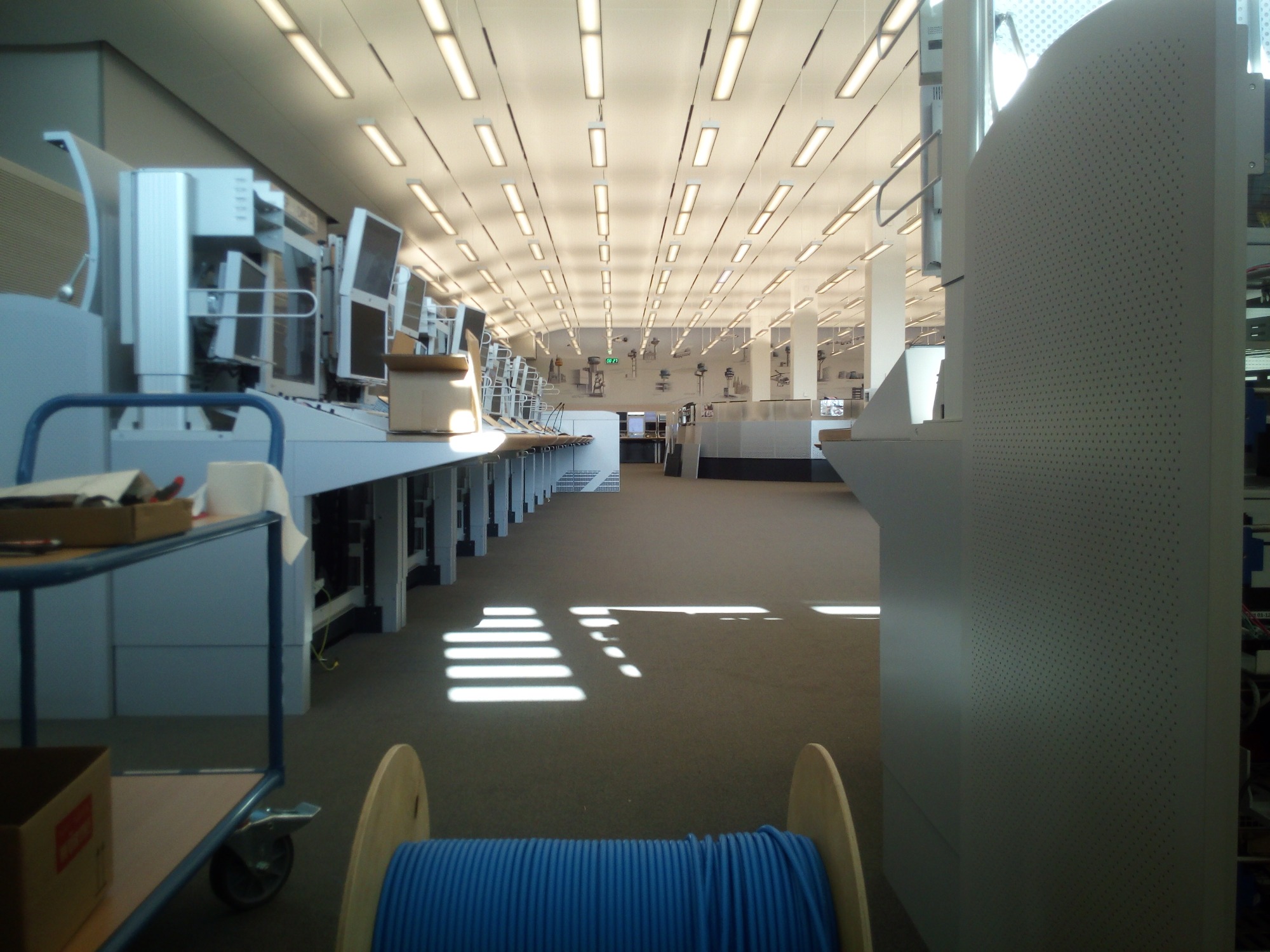 Długa sala z rzędem stanowisk komputerowych, rolka niebieskiego kabla na pierwszym planie, wpadające przez okno promienie słoneczne tworzą wzór na podłodze.