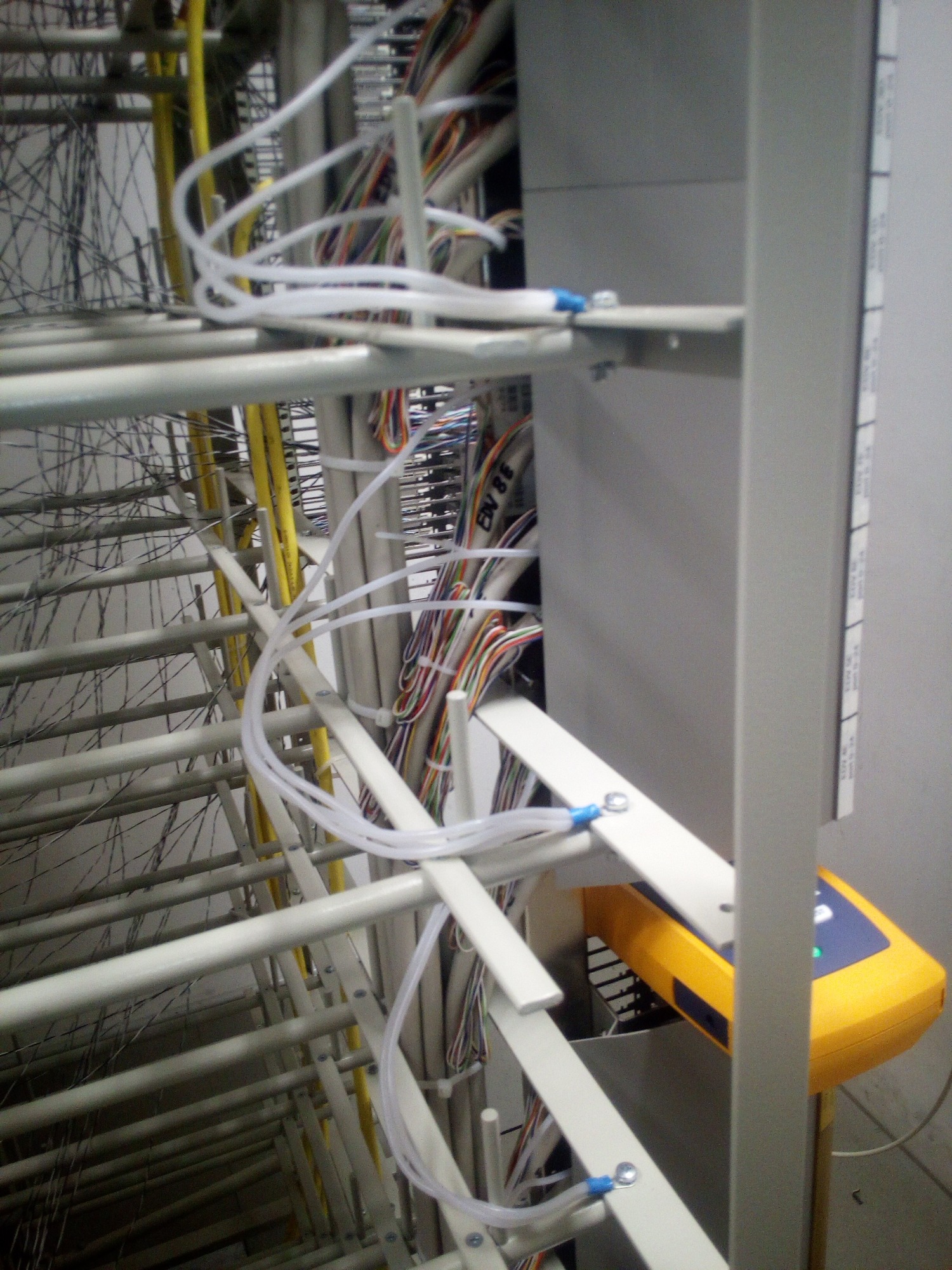 Szafa krosownicza z kablami telekomunikacyjnymi, z widocznym testerem kabli w kolorze żółtym i niebieskim, oraz uporządkowane przewody wewnątrz szafy.