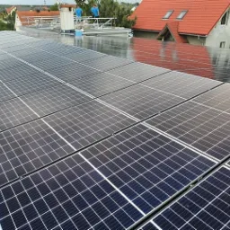 Instalacja paneli słonecznych na dachu domu jednorodzinnego, widoczni dwaj instalatorzy w niebieskich koszulkach, jasne niebo.