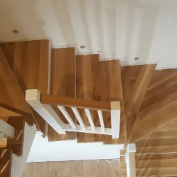 Widok z góry na drewniane schody ze stopniami w naturalnym odcieniu drewna i białą balustradą, z widocznymi otworami na oświetlenie wzdłuż ściany.