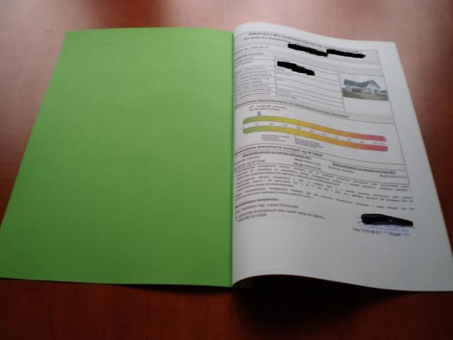 Otwarty segregator z zieloną okładką, prezentujący dokument z tabelami, wykresami i zdjęciem domu, prawdopodobnie dotyczącymi charakterystyki energetycznej budynku.