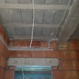 Surowy stan wnętrza budynku w trakcie budowy, widoczne ściany z czerwonej cegły, betonowy strop i instalacja elektryczna poprowadzona po ścianach i suficie.