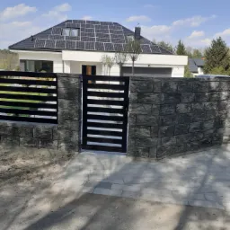Nowoczesna brama wjazdowa z poziomymi przęsłami, osadzona w murze z szarej kostki, na tle domu z panelami słonecznymi.
