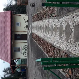 Ogród z domkiem: ścieżka z jasnych kamyków i romboidalnych płyt prowadzi do domu z czerwoną dachówką, ozdobionego dekoracyjnymi rowerami.