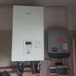 Biała pompa ciepła Panasonic Aquarea z panelem sterowania i manometrem, obok szary falownik Fronius Symo, rury instalacyjne na ścianie z jasnych płytek.