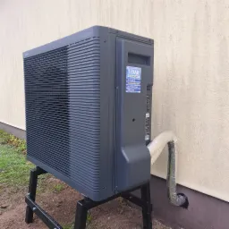 Szara pompa ciepła Panasonic Aquarea na czarnym stelażu, z izolowaną rurą przy ścianie budynku. Widoczna naklejka firmy instalacyjnej Libar.