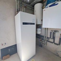 LIBAR Radosław Olbrych - Biała pompa ciepła Panasonic Aquarea Smart Cloud z zbiornikiem i instalacją hydrauliczną na tle surowej ściany. Widoczne rury i elementy montażowe.