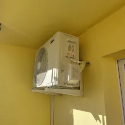 Biała jednostka zewnętrzna klimatyzatora marki AUX zamontowana na żółtej elewacji budynku, widoczne kable w osłonie i fragment okna po prawej stronie.