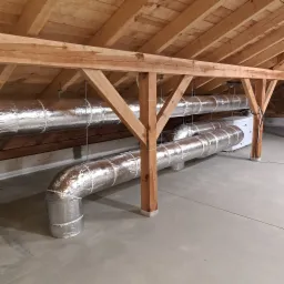 Widok z dołu na izolowane kanały wentylacyjne, podtrzymywane przez drewnianą konstrukcję więźby dachowej na poddaszu, z widocznym centralnym urządzeniem wentylacyjnym.