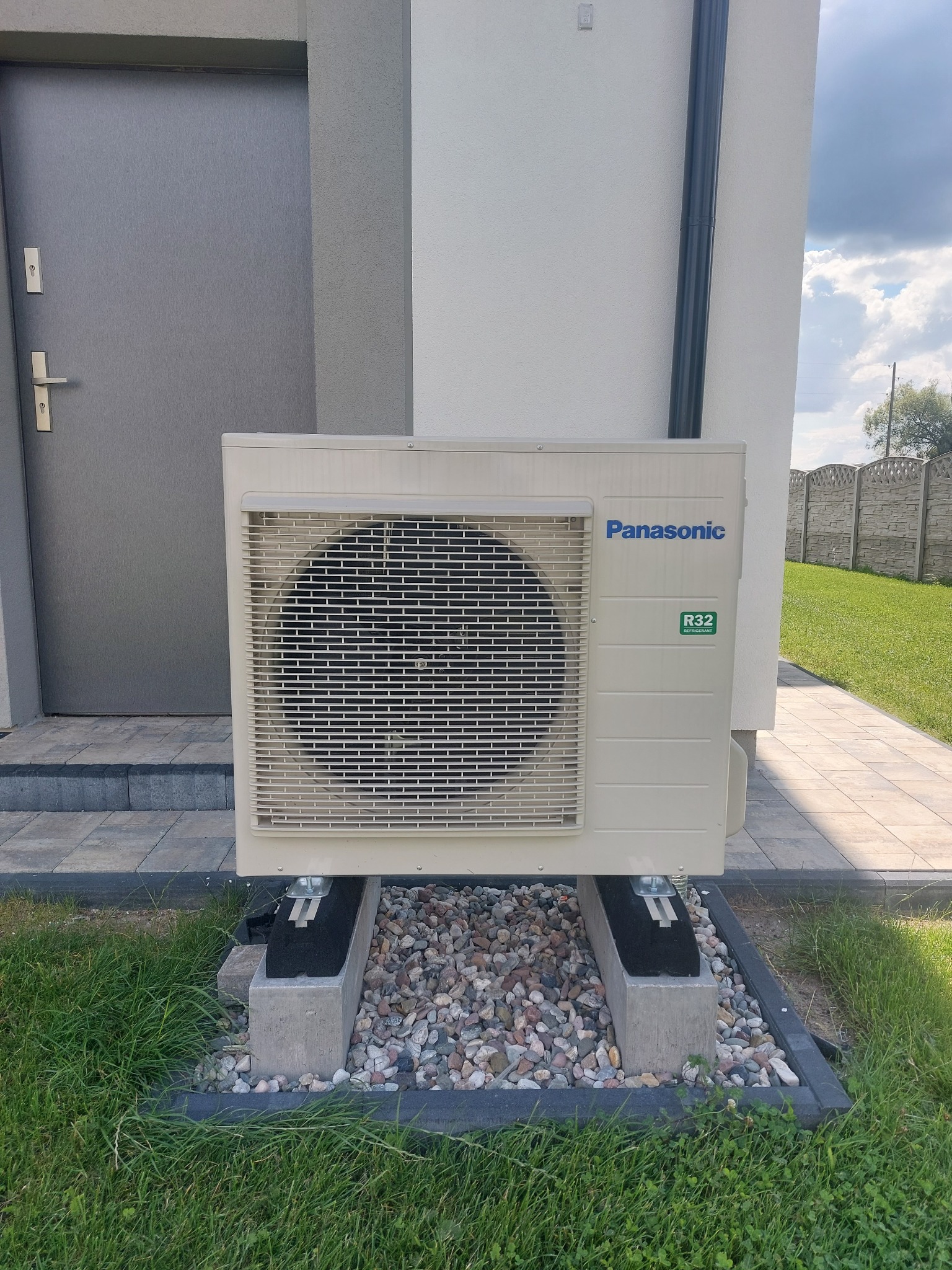 Zewnętrzna jednostka klimatyzacji Panasonic R32, zamontowana na betonowych wspornikach z podsypką z kamyków, obok wejścia do domu.