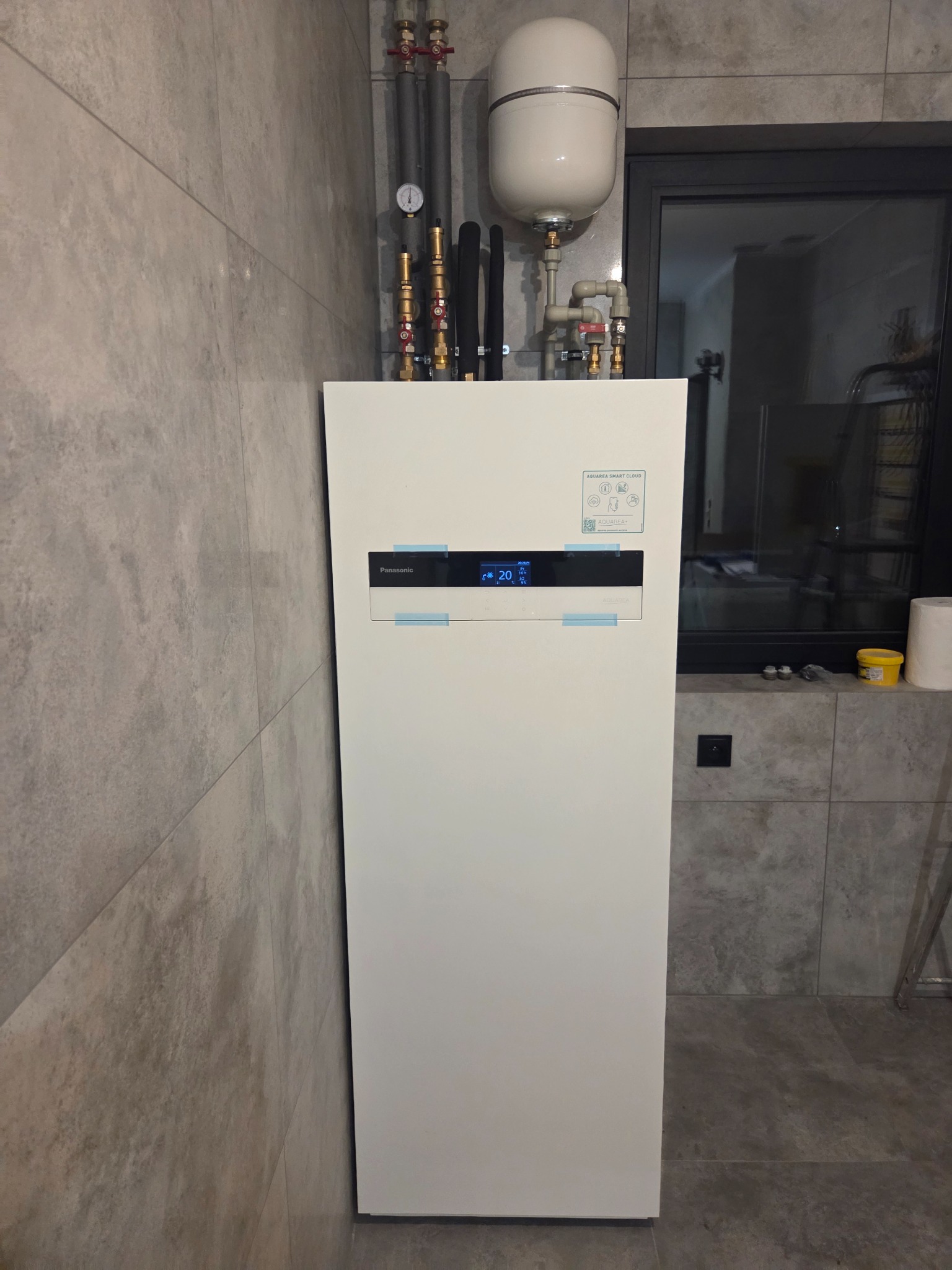 Biała pompa ciepła Panasonic Aquarea Smart Cloud z widocznymi rurami instalacji, manometrem i zbiornikiem wyrównawczym na tle szarej, płytkowej ściany i podłogi.