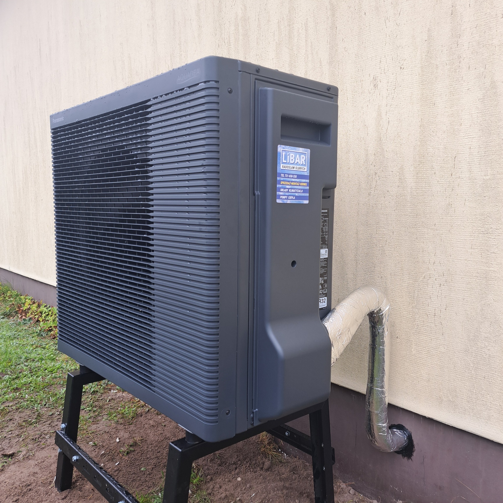Szara pompa ciepła Panasonic Aquarea na czarnym stelażu, z izolowaną rurą przy ścianie budynku. Widoczna naklejka firmy instalacyjnej Libar.