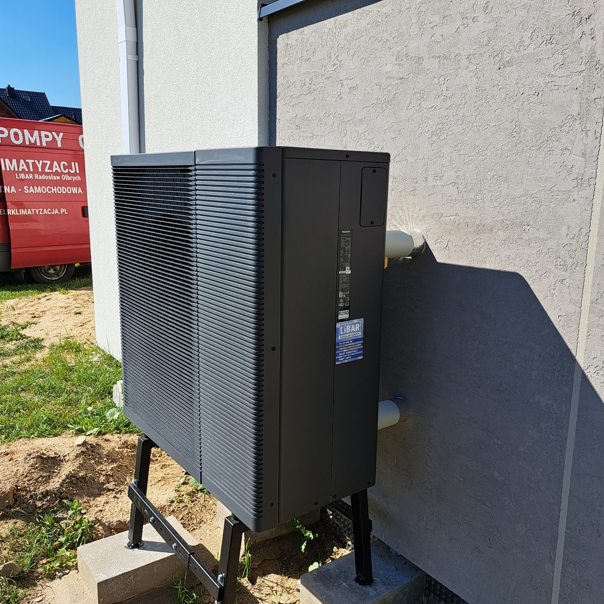 Czarna pompa ciepła Panasonic zamontowana na zewnątrz budynku, widoczna tabliczka firmy Libar. Ściana budynku o fakturze baranka, rury odprowadzające.
