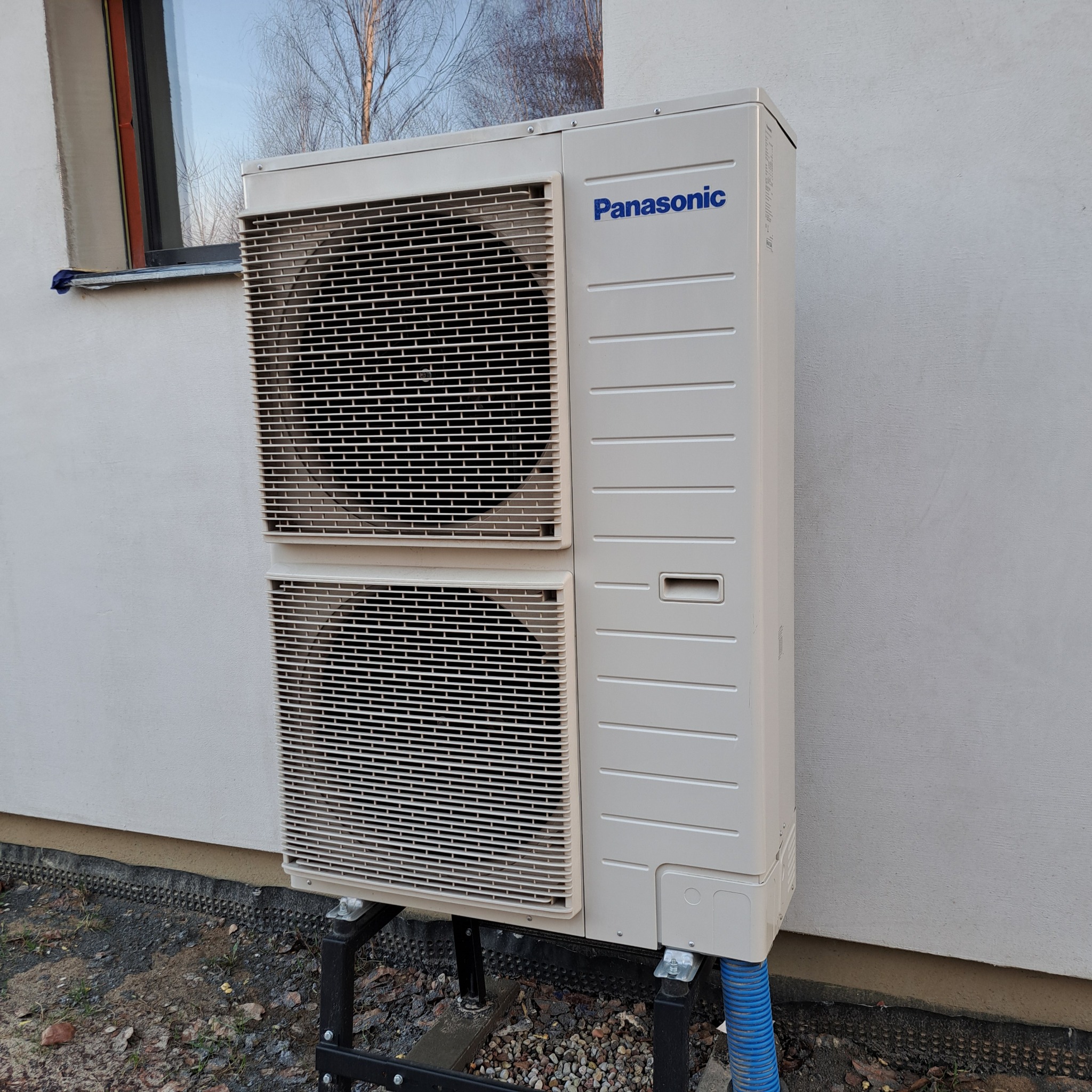 Zewnętrzna jednostka pompy ciepła Panasonic, zamontowana na czarnej, metalowej konstrukcji przy ścianie budynku. Widoczne niebieskie rury odprowadzające wodę.