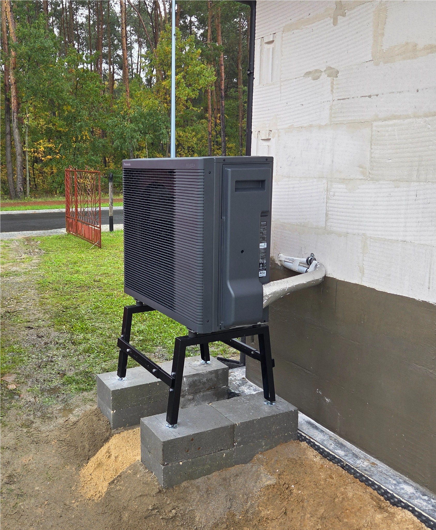 Zewnętrzna jednostka pompy ciepła Panasonic Aquarea na czarnym stelażu, ustawiona na betonowych bloczkach przy ścianie budynku w trakcie montażu. Widoczne rury.