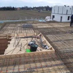 Zbrojenie stropu na budowie domu jednorodzinnego, widoczne szalunki, elementy zbrojeniowe, bloczki betonowe, w tle budynek w stanie surowym otwartym i pole uprawne.