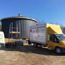 Cylindryczny zbiornik z blachy falistej w trakcie ocieplania, obok zaparkowany żółty samochód dostawczy z reklamą izolacji natryskowych oraz materiały budowlane i rusztowanie.