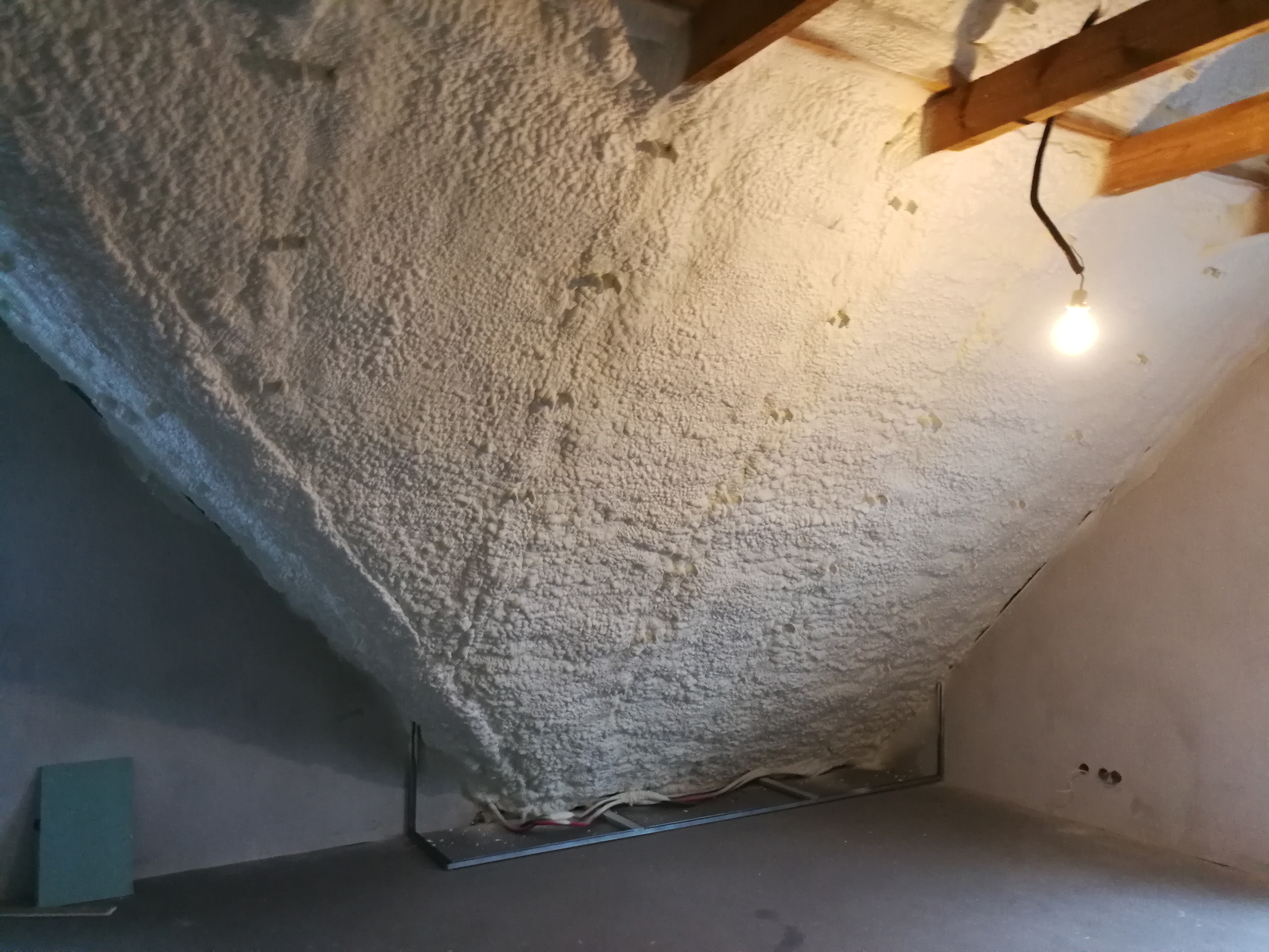 Poddasze po ociepleniu pianką PUR, widoczne drewniane belki stropowe i wystająca żarówka na kablu, na podłodze resztki materiałów montażowych.
