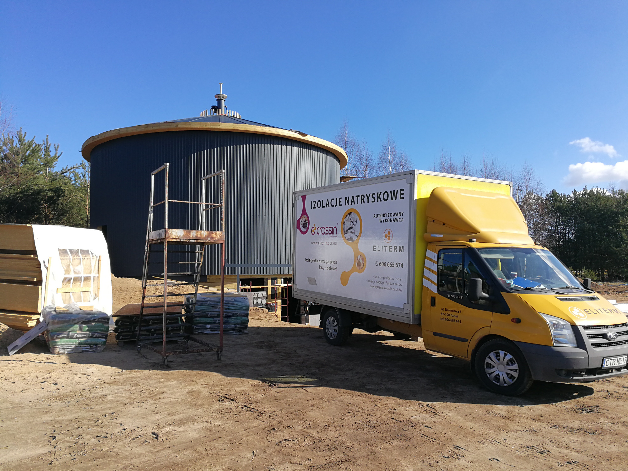 Cylindryczny zbiornik z blachy falistej w trakcie ocieplania, obok zaparkowany żółty samochód dostawczy z reklamą izolacji natryskowych oraz materiały budowlane i rusztowanie.