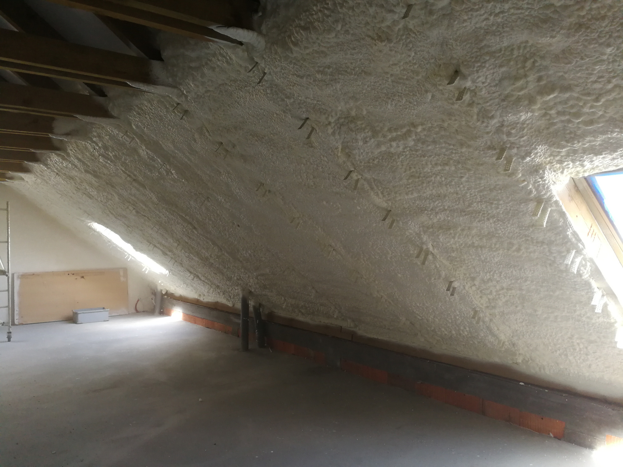 Poddasze z widoczną warstwą izolacji natryskowej, drewnianą konstrukcją dachu i oknem dachowym z widocznym niebieskim niebem.