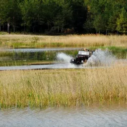 Ciemny jeep z otwartym dachem, w trakcie przejazdu przez płytkie jezioro, wzbijający fontanny wody, na tle trzcin i lasu.