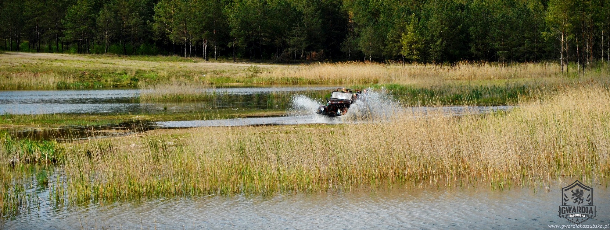 Ciemny jeep z otwartym dachem, w trakcie przejazdu przez płytkie jezioro, wzbijający fontanny wody, na tle trzcin i lasu.