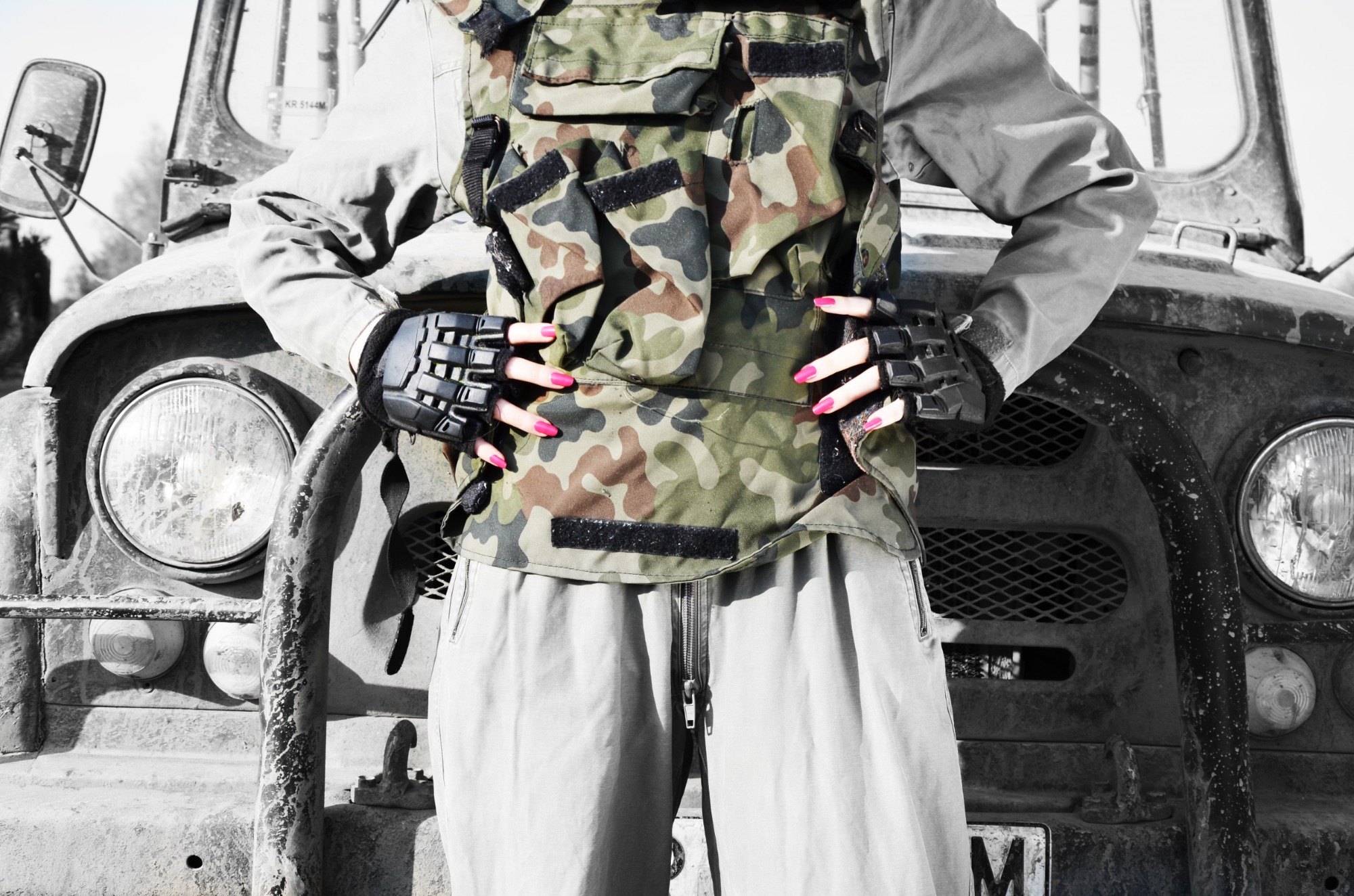 Kobieta w kamizelce moro i rękawicach taktycznych na tle starego, zardzewiałego samochodu terenowego z widocznymi reflektorami i grillem.