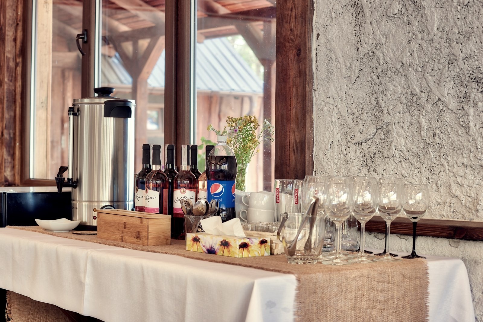 Stół z białym obrusem i jutowym bieżnikiem, przygotowany na przyjęcie: w tle okno, dystrybutor do napojów, butelki wina Carl Rossi, napój gazowany, szklanki, sztućce, serwetki w pudełku z motywem...