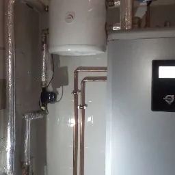 Instalacja grzewcza w kotłowni: biały zbiornik buforowy, niebieskie naczynie wzbiorcze, miedziane rury, zawory i izolowane termicznie przewody, widoczne elementy automatyki.