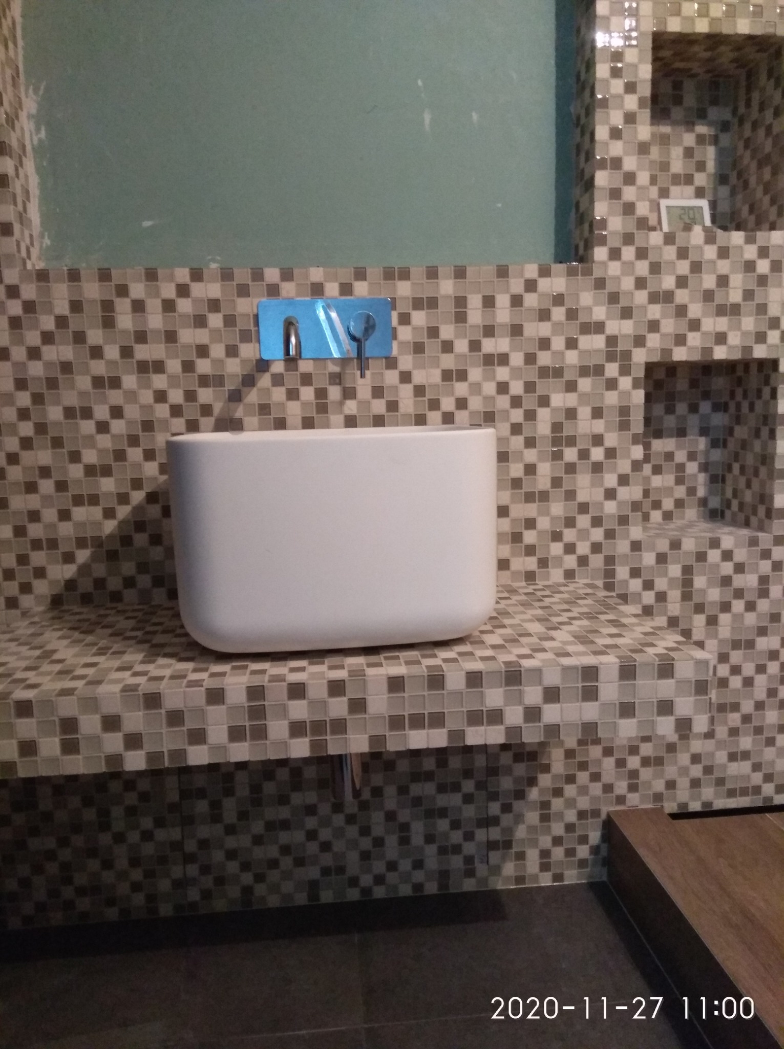 Nowoczesna, biała umywalka nablatowa o zaokrąglonych kształtach, zamontowana na blacie wyłożonym mozaiką w odcieniach szarości i beżu, z baterią podtynkową i fragmentem ściany z niedokończonym...