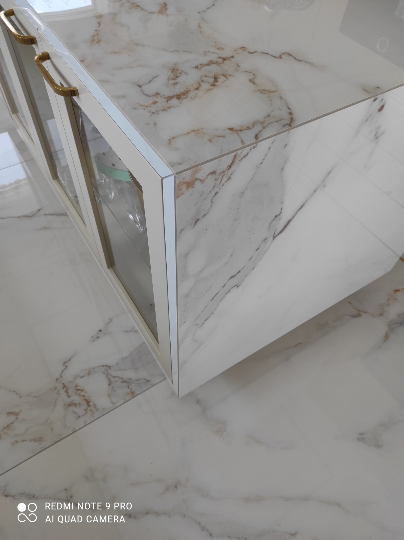 Biała szafka z marmurowym wzorem na blacie i frontach, złote uchwyty, widoczne szklane półki wewnątrz, dopasowana marmurowa posadzka.