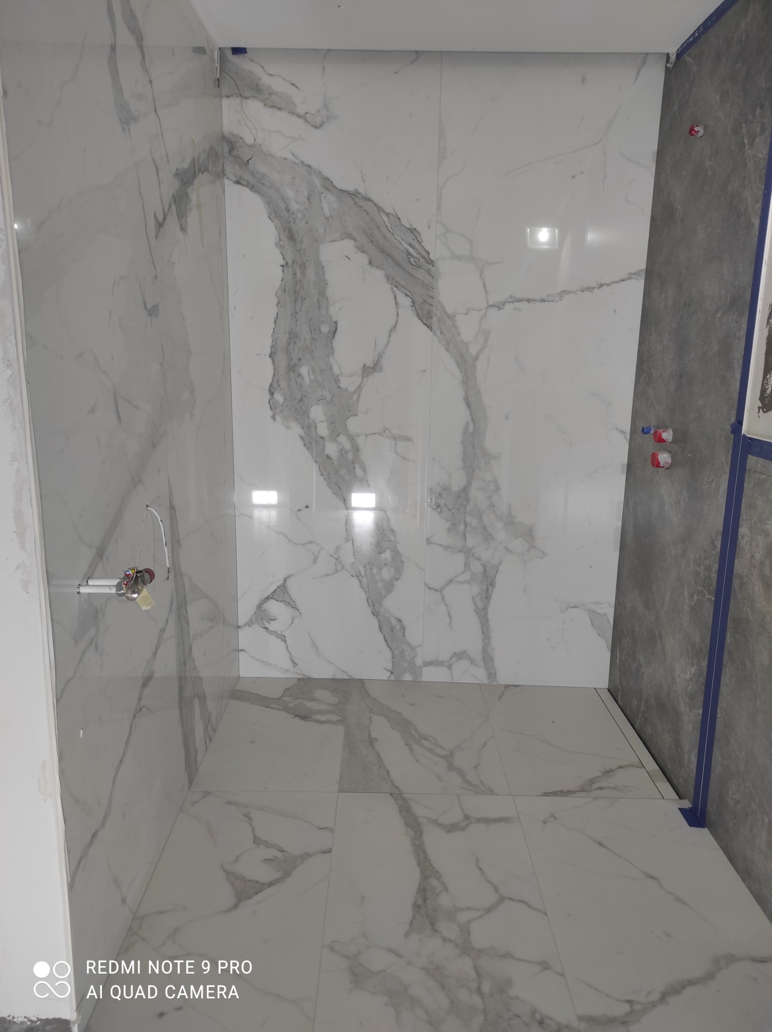 Wnętrze łazienki w trakcie wykańczania, widoczne białe płytki imitujące marmur na ścianach i podłodze, częściowo położone, z widocznymi instalacjami hydraulicznymi i fragmentem ściany przygotowanej...