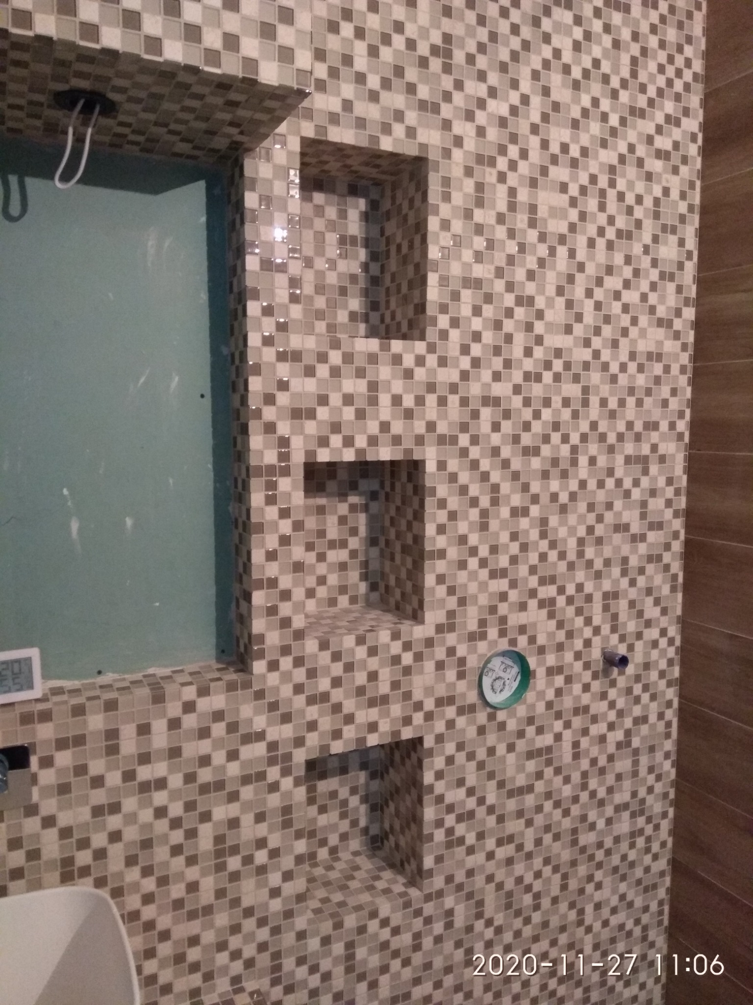 Ściana w łazience wyłożona mozaiką w odcieniach szarości i beżu, z wnękami wykończonymi tą samą mozaiką, widoczny fragment surowej ściany przygotowanej pod dalsze prace, w tle widoczny fragment...