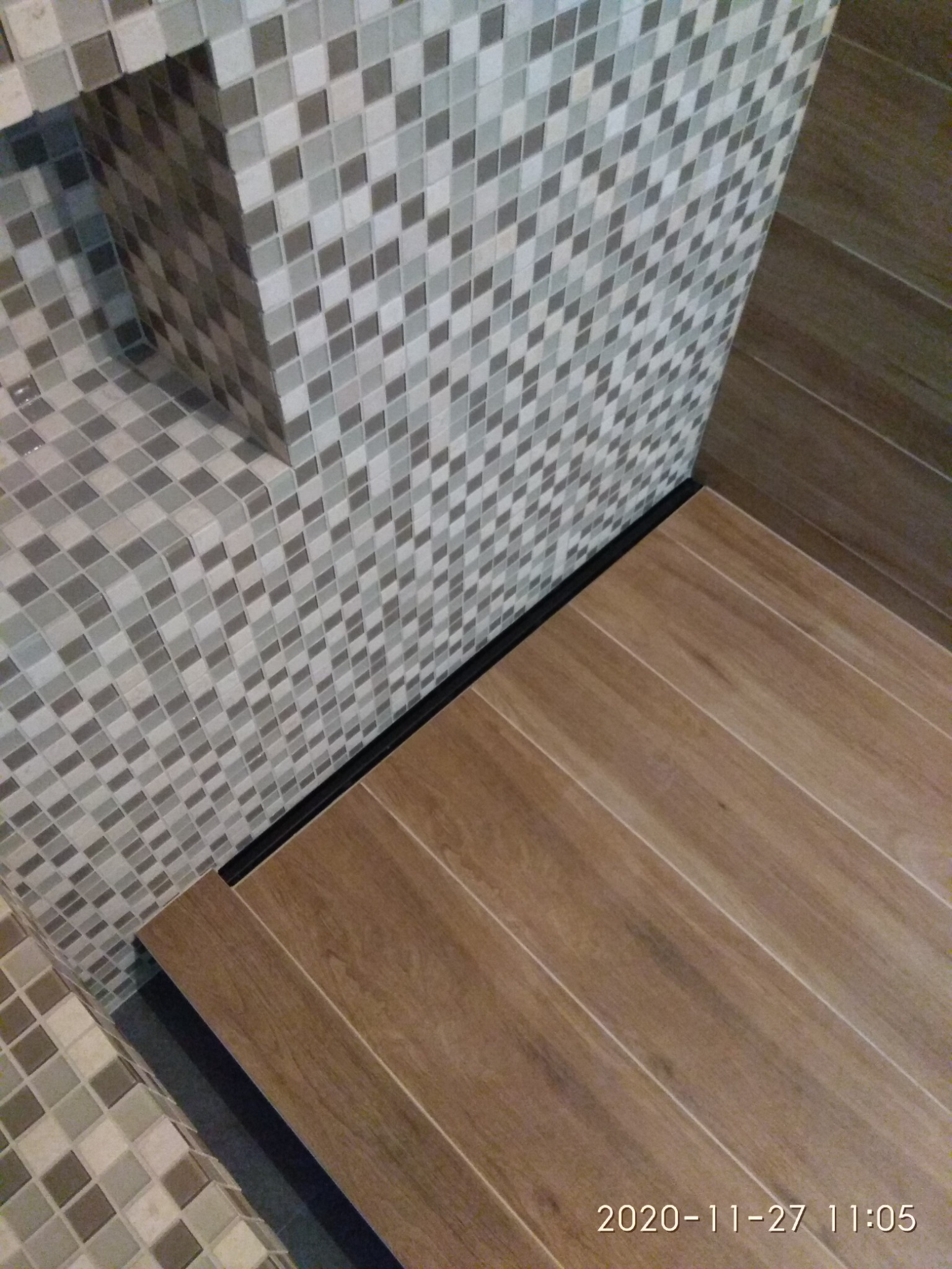 Wykończenie łazienki: mozaika w odcieniach szarości i beżu na ścianach oraz podłoga z płytek imitujących drewno, oddzielone czarną listwą.