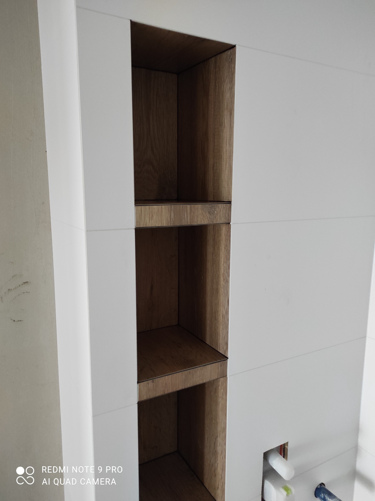 Biała ściana z trzema wnękami wykończonymi drewnem w kolorze orzechowym, widoczne fragmenty instalacji hydraulicznej w dolnej części ściany.