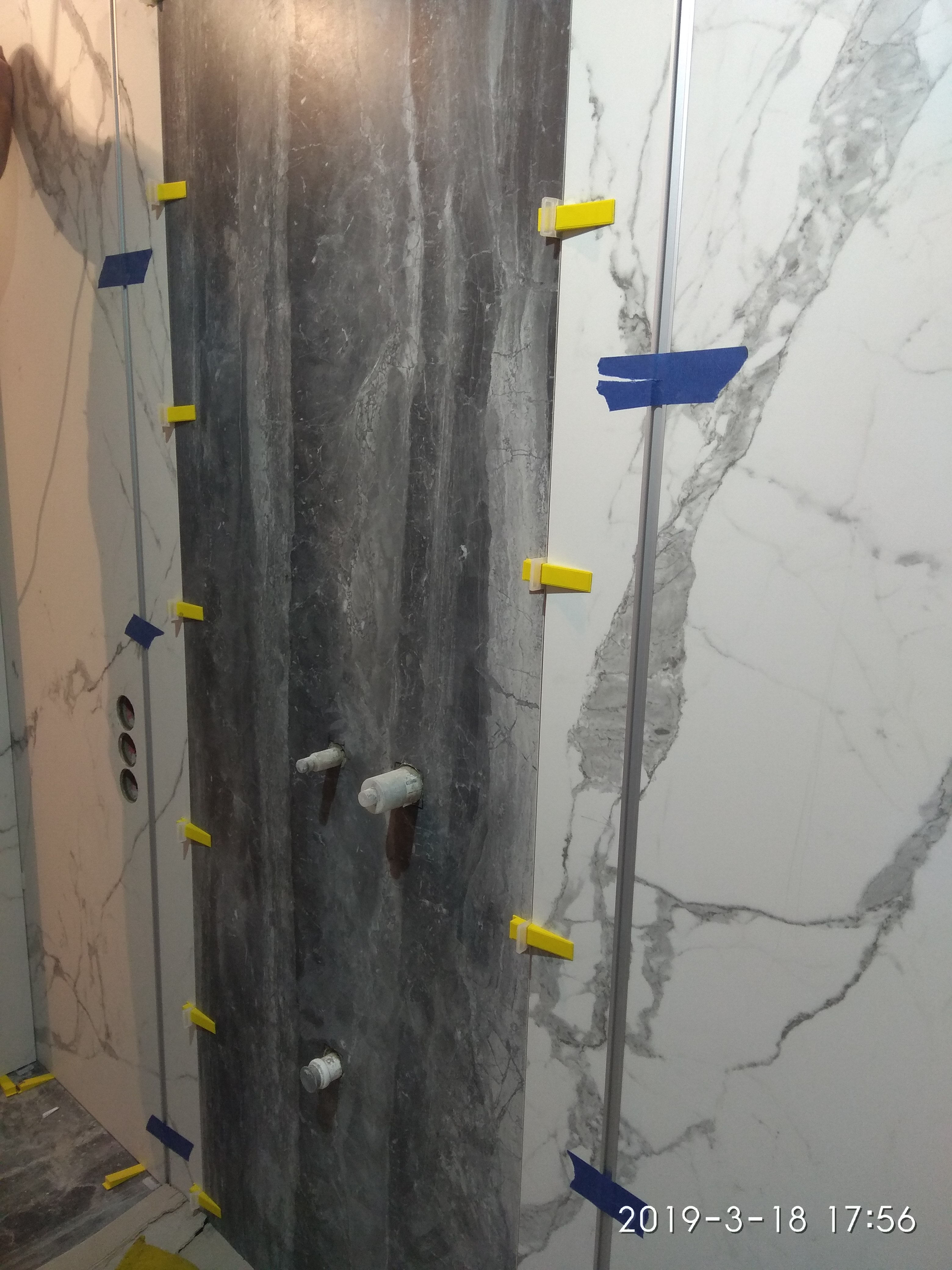 Montaż płytek imitujących marmur w łazience, widoczne żółte kliny dystansowe i niebieska taśma maskująca, przygotowane podejścia wodne.