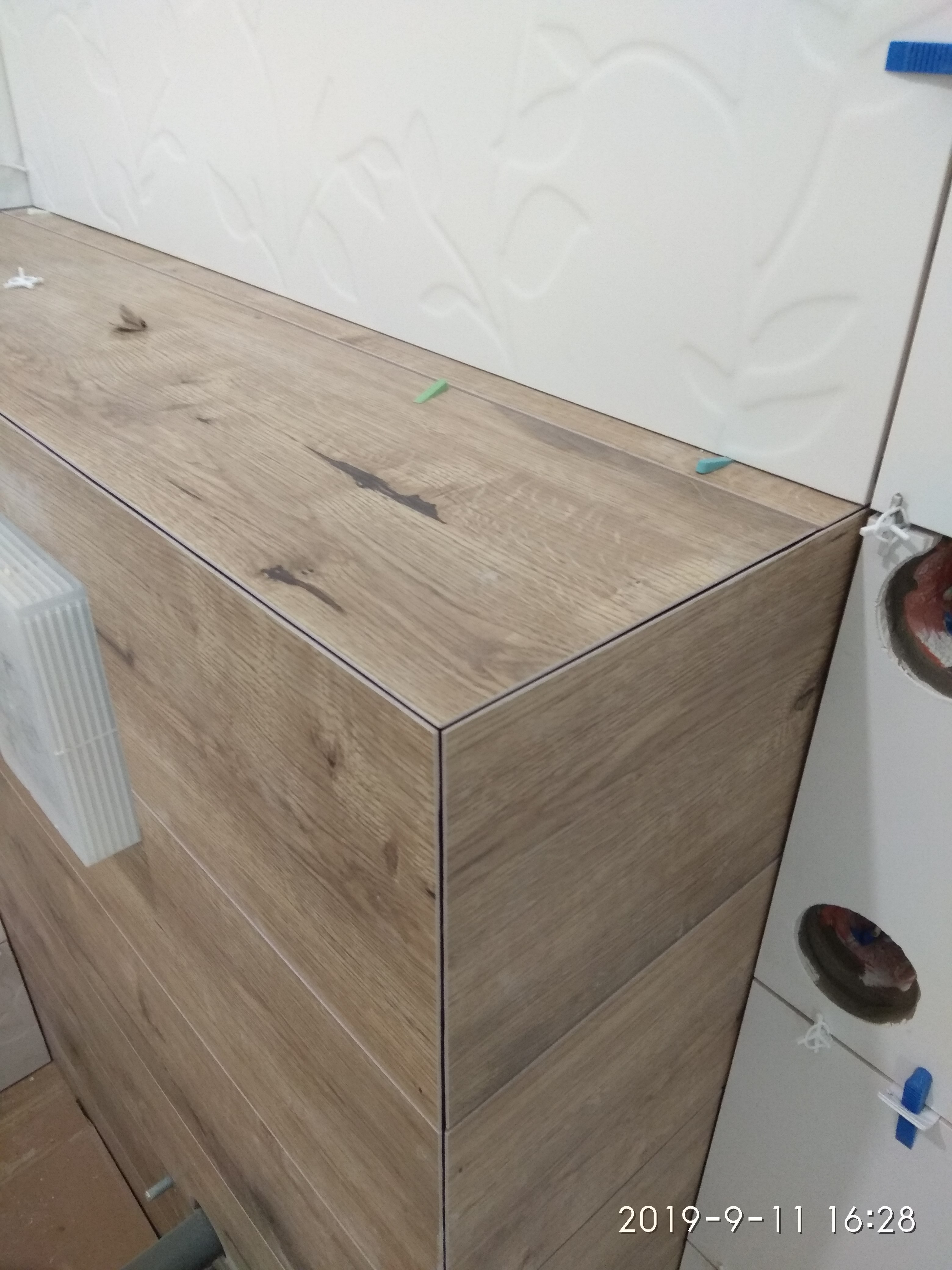 Wykończenie obudowy wanny płytkami imitującymi drewno, z widocznymi krzyżykami dystansowymi i białymi płytkami ściennymi z delikatnym wzorem.