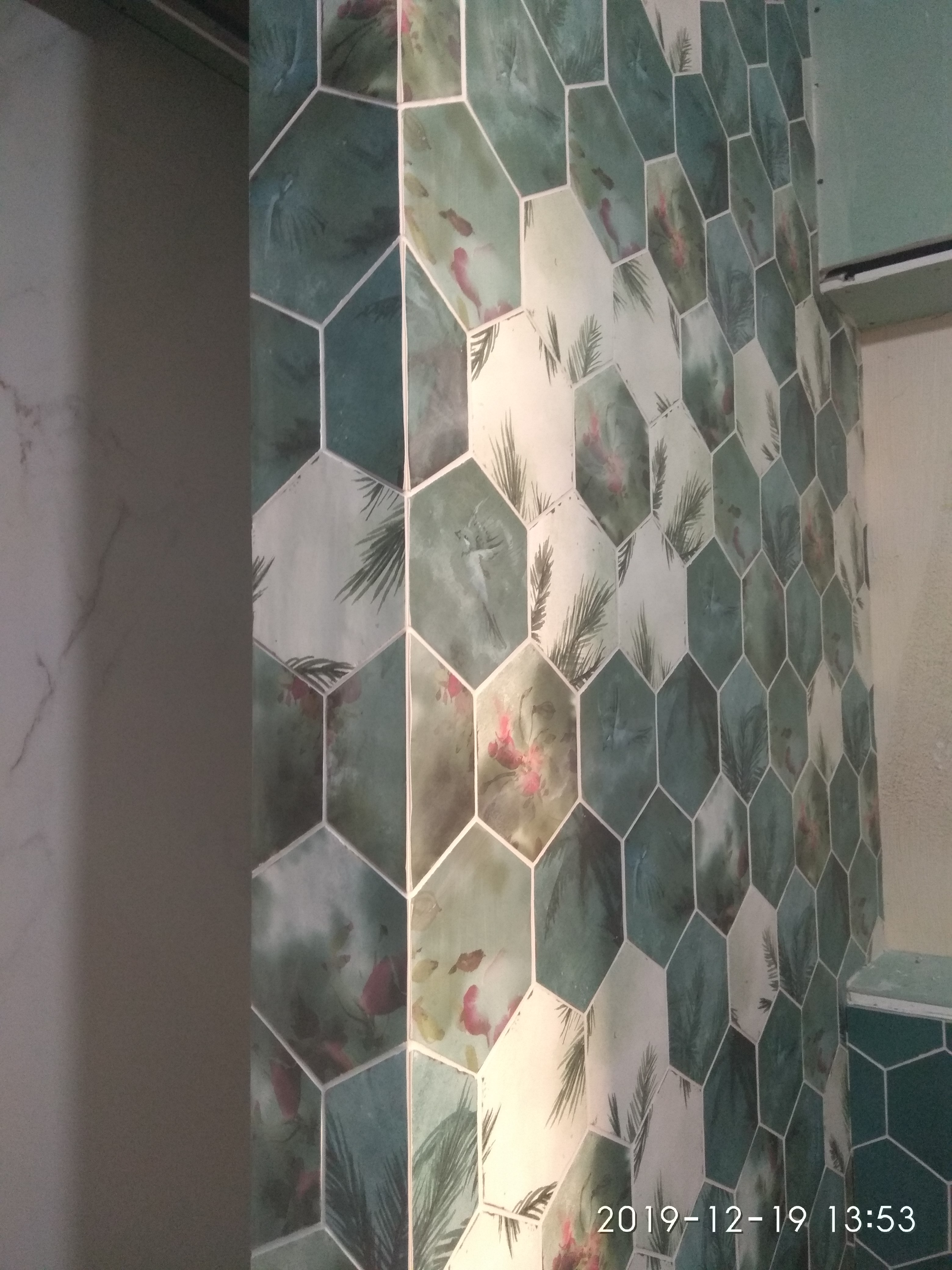 Ściana wyłożona heksagonalnymi płytkami z motywem tropikalnym, widoczne palmy i kwiaty w odcieniach zieleni i bieli, ułożone na ścianie w łazience.